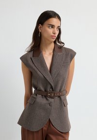 Kvinde med langt mørkt hår iført ærmeløs brun herringbone blazer med læderkrave og bælte, parret med matchende brune bukser.