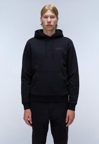 Zwarte hoodie met kangoeroezak, textuurstof en minimalistisch logodedetail op de borst. Draagt met zwarte broek.