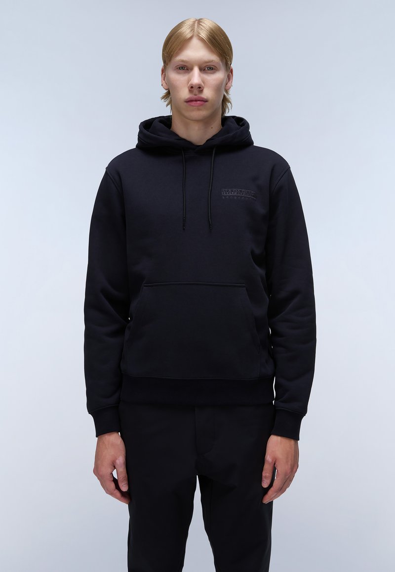 Zwarte hoodie met kangoeroezak, textuurstof en minimalistisch logodedetail op de borst. Draagt met zwarte broek.