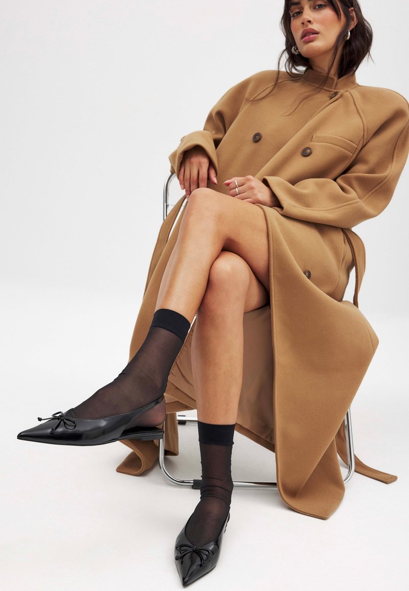 Manteau oversize marron avec des boutons double, associé à des ballerines noires pointues avec des nœuds et des chaussettes noires transparentes, assis sur une chaise métallique.