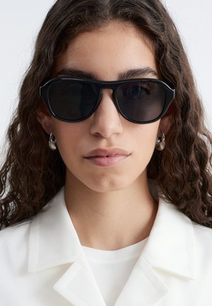 Mujer con cabello rizado y castaño, que lleva gafas de sol negras, pendientes de aro plateados y una chaqueta blanca con cuello, sobre un fondo liso.