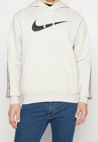 Man som bär en vit Nike-hoodie med svart logotyp och dragsko, i kombination med blå jeans, beskuren från axlarna till höfterna.