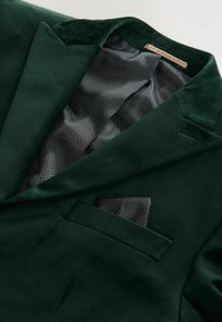 Ciemnozielony aksamitny żakiet z wyciętym kołnierzem, czarną siateczkową podszewką i małą poszewką w fakturowanym wzorze. Eleganckie detale projektu.