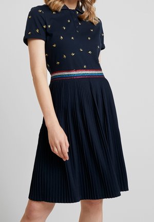 Kobieta ubrana w granatową sukienkę z krótkim rękawem, ozdobioną małymi wzorami żółtych ptaków, plisowaną spódnicą i wielokolorowym paskiem w paski.