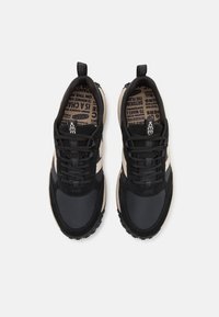 Schwarze und beige Sportschuhe mit einer Mischung aus Texturen, runden Schnürsenkeln und einer gemusterten Einlegesohle. Robuste Gummisohle mit Profil.