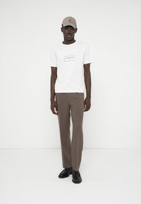 Biely tričko s čiernym logom, hnedé nohavice a čierne topánky. Taktiež nosí béžovú čiapku. Hladká látka a priliehavý strih.