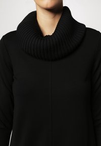 Czarny sweter z grubym, prążkowanym wysokim kołnierzem i gładką, dopasowaną sylwetką. Materiał wydaje się miękki i ma minimalistyczny design.