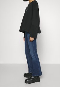 Person som bär en lös svart långärmad tröja, blå utsvängda jeans och svarta klumpiga platåstövlar, som står mot en enkel vit bakgrund.