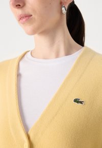 Žlutý pletený svetr s výstřihem do V, texturovaná látka a zelený vyšitý krokodýlí logo na levé straně. Pod ním bílá žebrovaná košile.