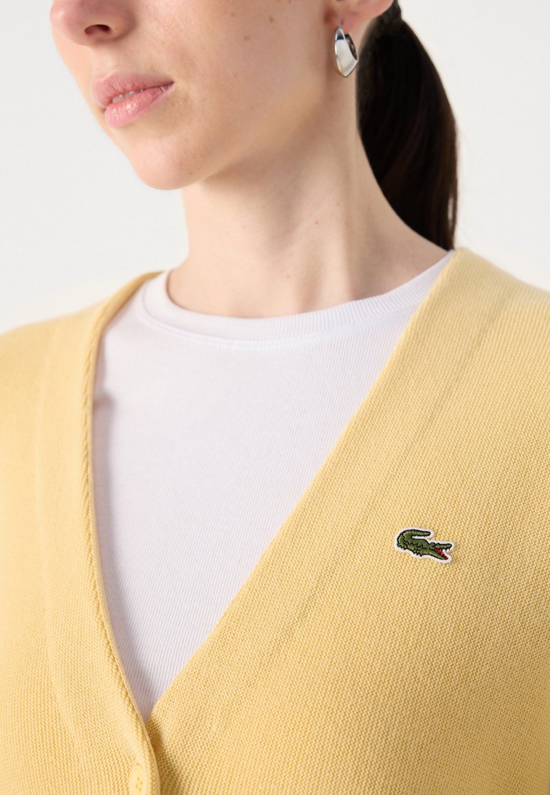 Gelber gestrickter Cardigan mit V-Ausschnitt, strukturiertem Stoff und grünem gesticktem Krokodil-Logo auf der linken Seite. Darunter ein weißes geripptes Shirt.