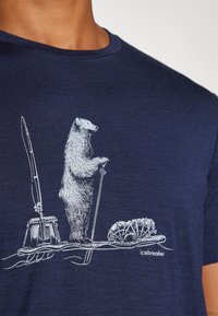 Camiseta azul marino con un gráfico blanco de un oso de pie con un remo, al lado de un trineo y un palo, sobre una tela texturizada.
