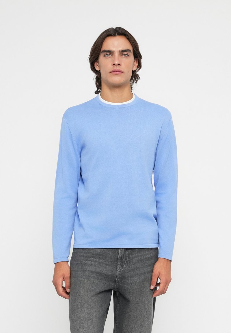 Suéter de manga longa azul claro em um tecido de malha suave com gola redonda, usado sobre jeans cinza escuro. Design simples e clássico.