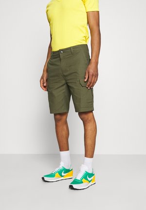 Muž nosící žluté tričko, olivově zelené cargo kraťasy, bílé ponožky a zelené, žluté a bílé tenisky Nike stojí proti jednobarevnému pozadí.