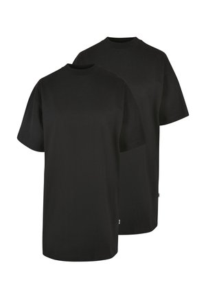 Femme aux cheveux bouclés portant une robe noire ample à manches courtes, debout sur un fond gris uni.