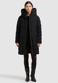 Melnā pūkains jaka ar kapuci, kas aprīkota ar rāvējslēdzēju un pogu aizdari, vertikālām virsmas gumijām, sānu kabatām un gludu, spīdīgu tekstūru.