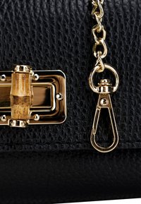 Sort, tekstureret lædertaske med guldhardware, bambus-toggle-lukning og guldkædestropp. Har en cirkulær låse detalje.