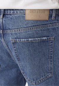 Siniset farkut, joissa on nahkainen tagi, jossa lukee "ONLY & SONS" takavatsassa, näkyvillä ompeleilla ja tavallisella taskusuunnittelulla.