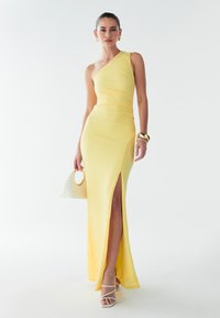 Asymmetrisches gelbes Kleid aus strukturiertem Stoff, mit seitlichem Schlitz und taillierter Silhouette. Kombiniert mit weißen Absatzsandalen und einer kleinen Tasche.