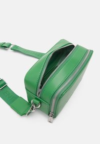 Zelená kožená taška přes tělo s dvěma stříbrnými zipy a nastavitelným popruhem, na kterém je embosováno jméno značky, zobrazená částečně otevřená na bílém pozadí.
