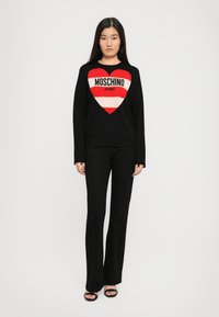 Svart hettegenser med et rødt og hvitt hjerte-design som har teksten "MOSCHINO JEANS"; kombinert med svarte vide bukser og åpen tå-sko.