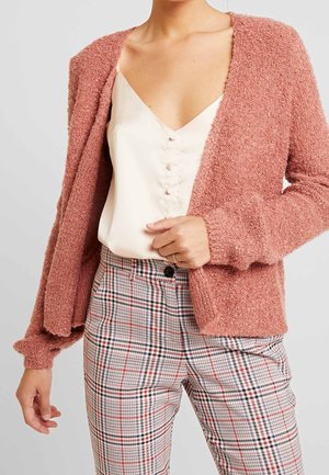 Femme portant un cardigan texturé rose poudré sur un chemisier blanc à boutons, avec un pantalon à carreaux taille haute dans des tons neutres.