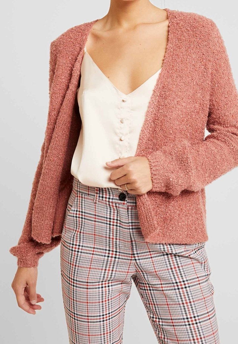 Femme portant un cardigan texturé rose poudré sur un chemisier blanc à boutons, avec un pantalon à carreaux taille haute dans des tons neutres.