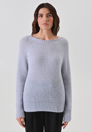 Donna con lunghi capelli scuri, vestita con un maglione lavorato a maglia grigio chiaro e pantaloni neri, in piedi di fronte a uno sfondo chiaro e semplice.