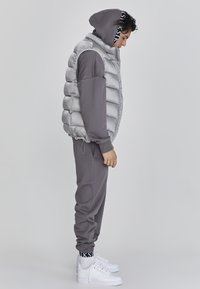 Sivi puffer prsluk s visokim ovratnikom, upareni s odgovarajućim sivim trenirkama. Bijele tenisice s crnim logotipom na manžetama. Glatke teksture kroz cijeli outfit.