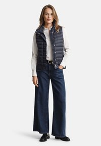 Tamsiai mėlyna pūkinė liemenė su aukštu apykakle, užsegama užtrauktuku ir spaustukais, dėvima ant balto marškinio, derinama su plačiomis džinsinėmis kelnėmis ir juodais batais.