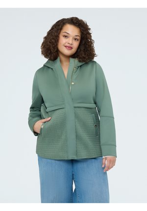 Žena s kudrnatými vlasy, oblečená v světle zelené texturované bundě a světle modrých džínách, stojící s jednou rukou v kapse na jednobarevném pozadí.