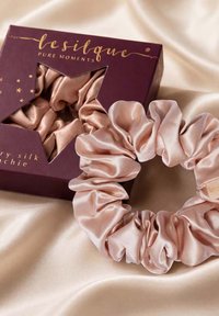 Chouchou en soie rose clair avec étiquette logo dorée à côté d'une boîte violette avec une fenêtre en forme d'étoile et texte doré sur tissu satin crème.
