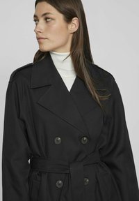 Juodas dvigubas lietpaltis su diržu, plačiais atvartu ir dviem dideliais mygtukais. Lygus audinys su švariu, pritaikytu dizainu.