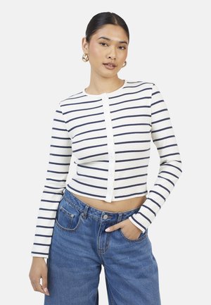 Kobieta o ciemnych włosach, w złotych kolczykach kółkach, ubrana w białą, krótko skrojoną koszulę na guziki z długim rękawem z granatowymi poziomymi paskami oraz niebieskie dżinsy.