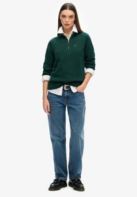 Groene gebreide trui met een rits, gedragen over een witte kleding met kraag. Gecombineerd met blauwe rechte jeans en zwarte schoenen.