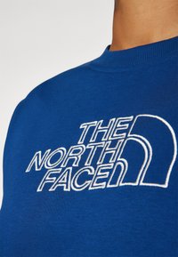 Kék pamut pulóver, fehér hímzett logóval, amely a "THE NORTH FACE" feliratot és ívelt díszítőelemeket tartalmaz. Puha tapintású, bordázott nyakkivágással.