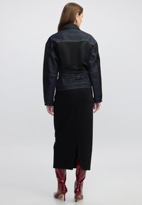 Giacca di denim con pannelli in satin neri, vita cintrata e cuciture a contrasto; abbinata a un vestito midi nero e stivali al ginocchio rossi lucidi.