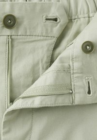 Pantalon vert clair avec braguette à boutons et fermeture éclair, montrant des boutonnières cousues et les détails de la texture du tissu.