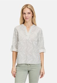 Blusa bianca con un motivo a piuma testurizzato, maniche 3/4 arrotolate, scollo a V e vestibilità ampia. Tessuto leggero e semitrasparente.