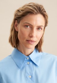 Camicia blu chiaro con colletto, fronte con bottoni e tessuto liscio. Presenta un design classico e una leggera lucentezza. Nessun motivo o dettaglio visibile.