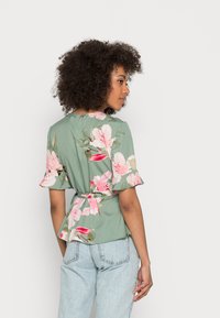 Blusa floral em tecido verde com flores rosa e brancas, mangas curtas com babados e detalhe na cintura amarrado. Usada com jeans azul claro.