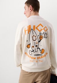 Krem sweatshirt med et lekent grafisk design som har en surfbrett-karakter, oransje tekst og tilpassbare detaljer på baksiden.