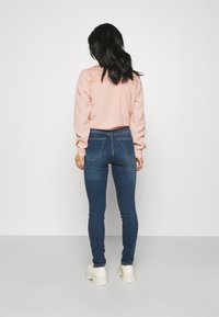 Sweatshirt rosa claro com detalhes em renda nos ombros, combinado com jeans skinny azul escuro e ténis brancos de plataforma. Vista traseira.