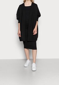 Woman wearing a black ribbed dress, loose black cardigan, and white platform sneakers, standing on a gray floor against a white wall.