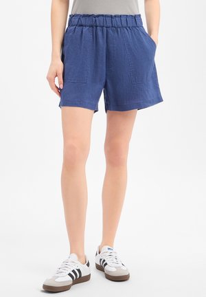Femme portant un short bleu à taille élastique avec poches plaquées à l'avant et des baskets blanches à rayures noires, debout sur fond blanc.