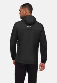 Homme de dos portant une veste à capuche Mammut noire et un pantalon noir devant un fond gris clair uni.