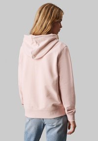 Rosa hettegenser laget av myk stoff, med kengurulomme, ribbede mansjetter og kant, og en avslappet silhuett.