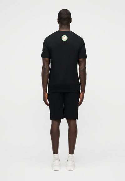 Άνδρας που στέκεται με την πλάτη γυρισμένη, φοράει μαύρο μπλουζάκι με κυκλικό λογότυπο κοντά στη γραμμή του λαιμού στο πίσω μέρος, μαύρο σορτς, άσπρες κάλτσες και άσπρα αθλητικά παπούτσια.