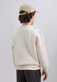 Krémová mikina s kulatým výstřihem, žebrovanými manžetami a lemem, v kombinaci s tmavými manšestrovými kalhotami. Čepice je béžová s nastavitelným páskem.