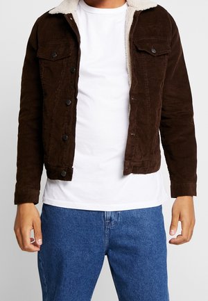 Hombre con chaqueta marrón de pana con cuello de borrego blanco, camiseta blanca y vaqueros azules, de pie contra un fondo claro y liso.