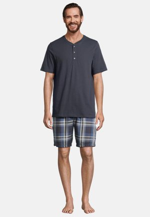 Marineblaue Kurzarm-Henley-Shirt mit drei Knöpfen, kombiniert mit karierten Shorts in Blau-, Grau- und Weißtönen. Barfuß-Positur.